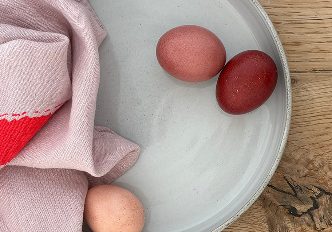 Du siehst einen Tisch, darauf eine Schale mit drei naturgefärbten Eiern und einem Geschirrtuch, ein Bild zum Artikel Eier natürlich färben mit Avocadoschalen – rosa und rote Naturfarbe aus Avocadokernen