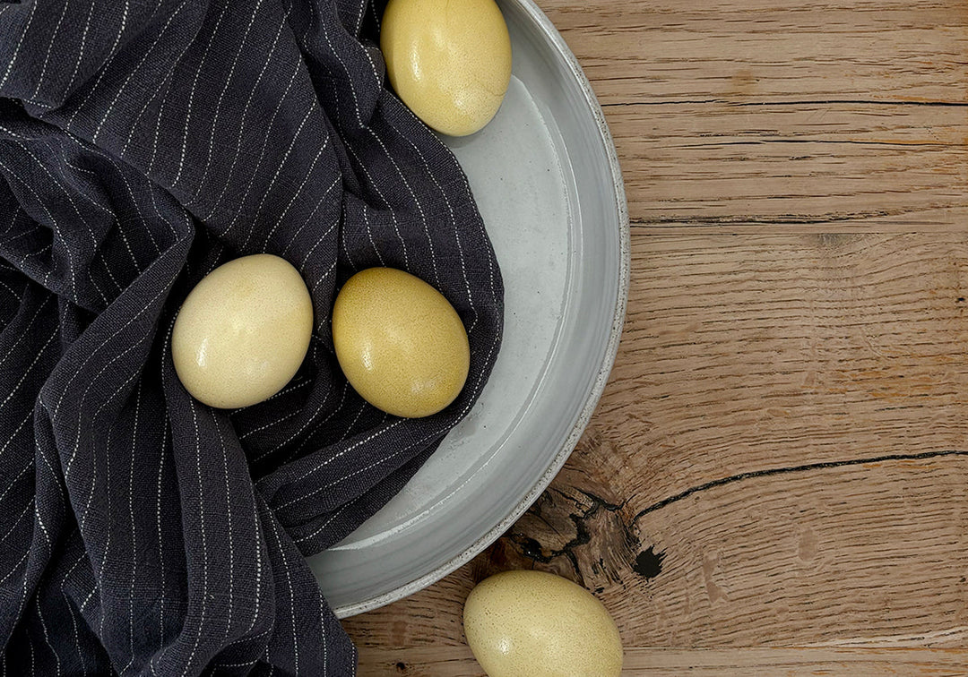 Du siehst einen Holztisch, darauf eine Keramikplatte und ein schwarz-weiss-gestreiftes Geschirrtuch sowie mit Kamillenblüten gefärbte Eier. Sanfte Gelbtöne aus Kamillenblüten