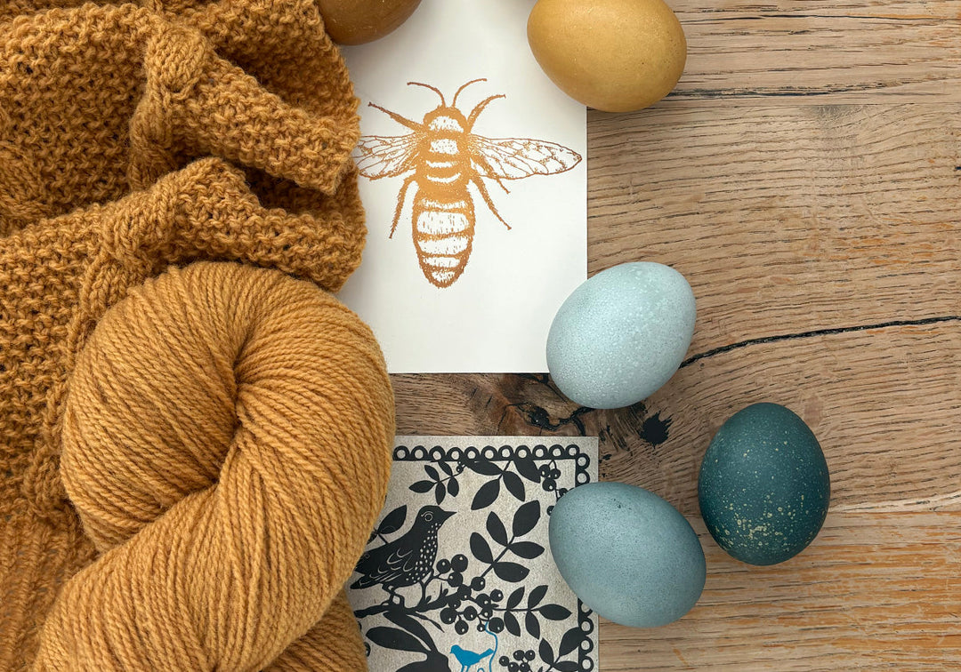 Du siehst einen Tisch mit gefärbter, verstrickter Wolle in der Farbe Honig (Rhabarberwurzeln), Postkarten, gefärbte Eier mit Granatapfelschalen und schwarzen Malvenblüten. Wie das geht erfährst Du in diesem Artikel.